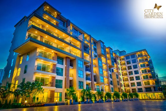 Modern apartment building illuminated at dusk with greenery and a blue sky.